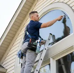 Professional window cleaner on a ladder using a squeegee to clean an exterior arched window, Erie, PA.